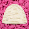 Vista completa gorro crochet crudo Ivory Heart con charm corazón perlado y placa LIA.G — fondo rosa