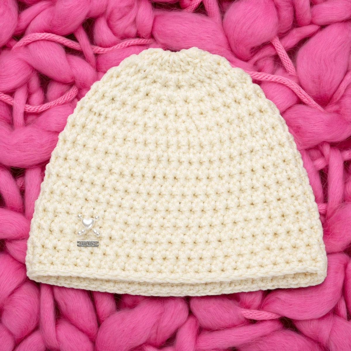 Vista completa gorro crochet crudo Ivory Heart con charm corazón perlado y placa LIA.G — fondo rosa