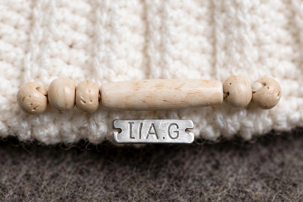 Detalle cuentas de madera natural ensartadas horizontalmente en puño y placa metálica LIA.G — Cream Cable Grey Pom