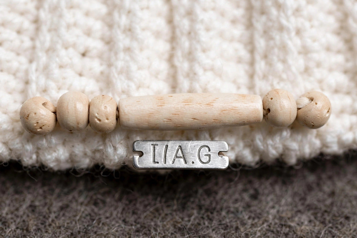 Detalle cuentas de madera natural ensartadas horizontalmente en puño y placa metálica LIA.G — Cream Cable Grey Pom